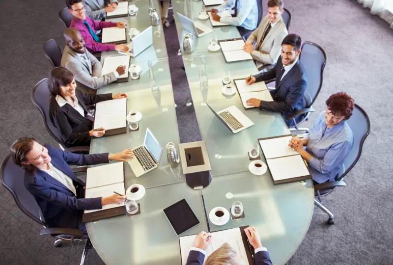 15 People sitting around a large conference table with laptops and documents, engaged in a meeting.