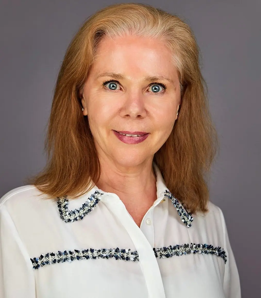 IMG_3414 A woman with shoulder-length hair, wearing a white blouse with decorative trim, stands against a plain gray background.