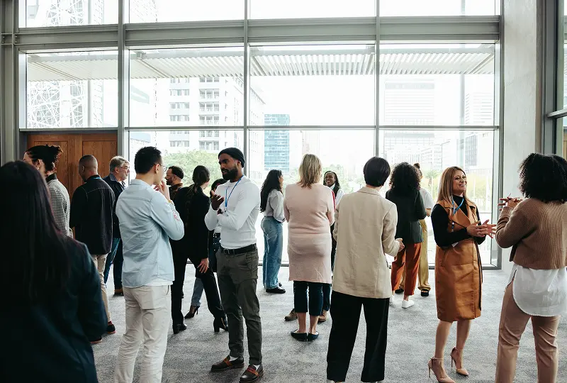 A group of people standing and conversing in a spacious room with large windows and a city view.